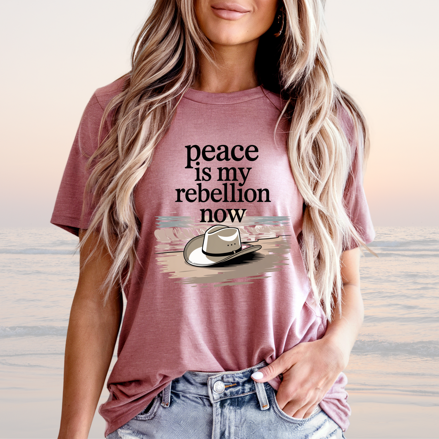 Woman wearing mauve Peace is My Rebellion Now Tee with cowboy hat graphic and loose fit by the beach