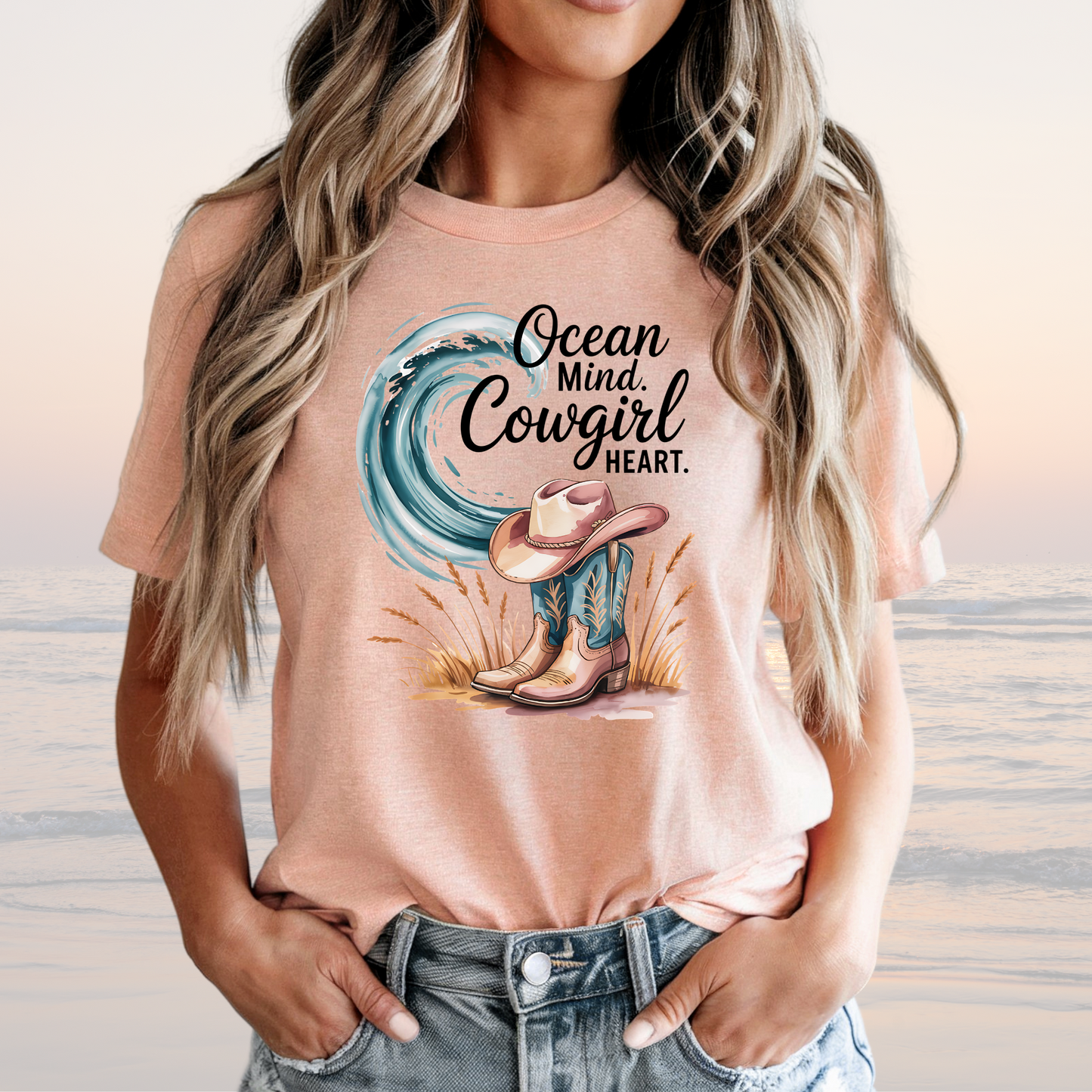 Woman wearing a peach Ocean Mind Cowgirl Heart Tee with wave, cowboy boots, and hat graphic, styled casually with jeans against a beach background