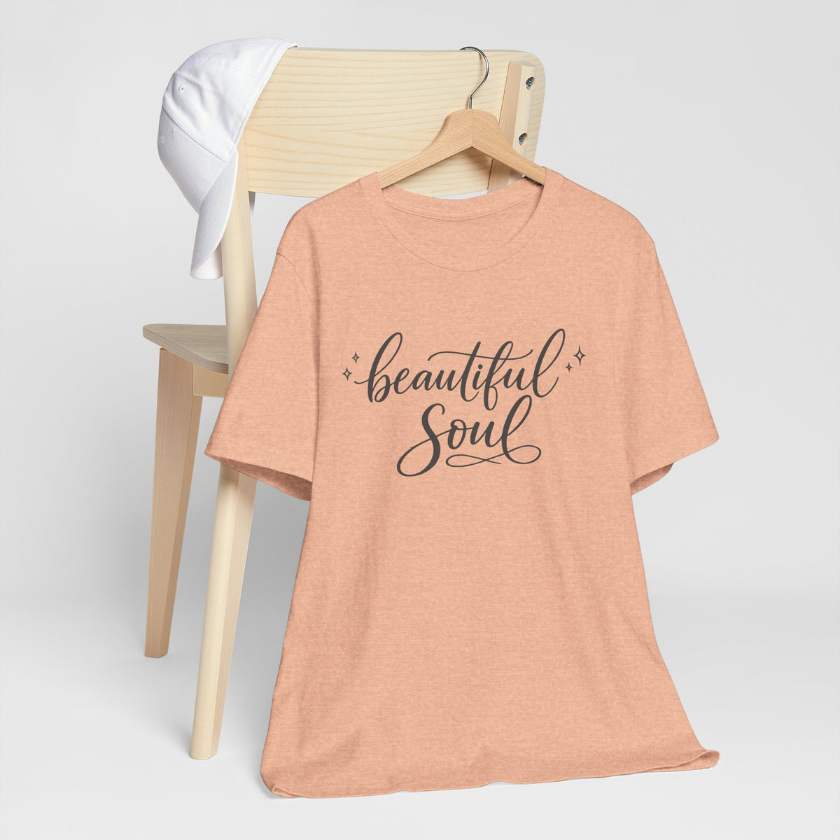 A peach-colored short-sleeve t-shirt with 'beautiful Soul' printed in black script on the front, hanging on a wooden hanger from a light wood chair with a white shirt draped over it, against a clean white background.