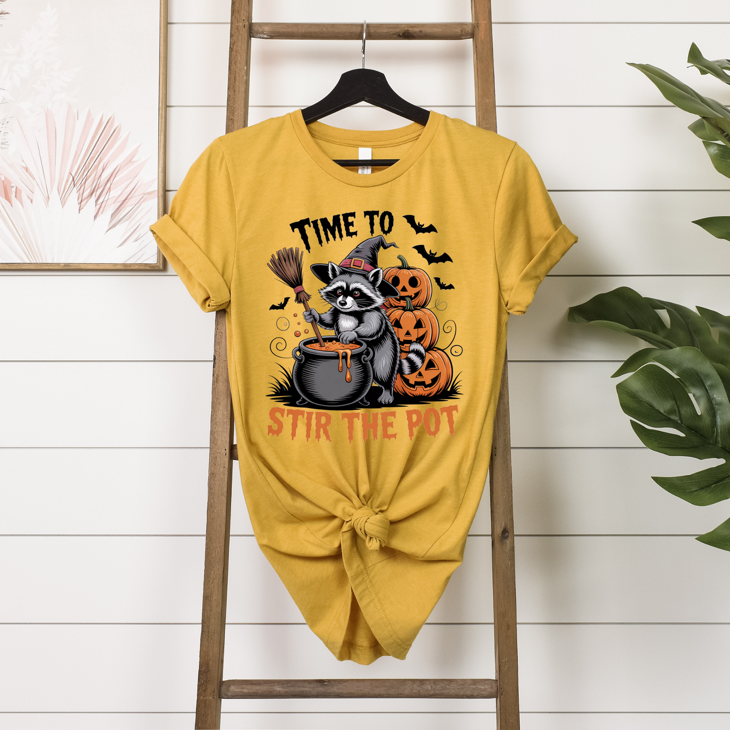 Yellow t-shirt with Halloween-themed design of a raccoon and text, hanging on a wooden ladder.