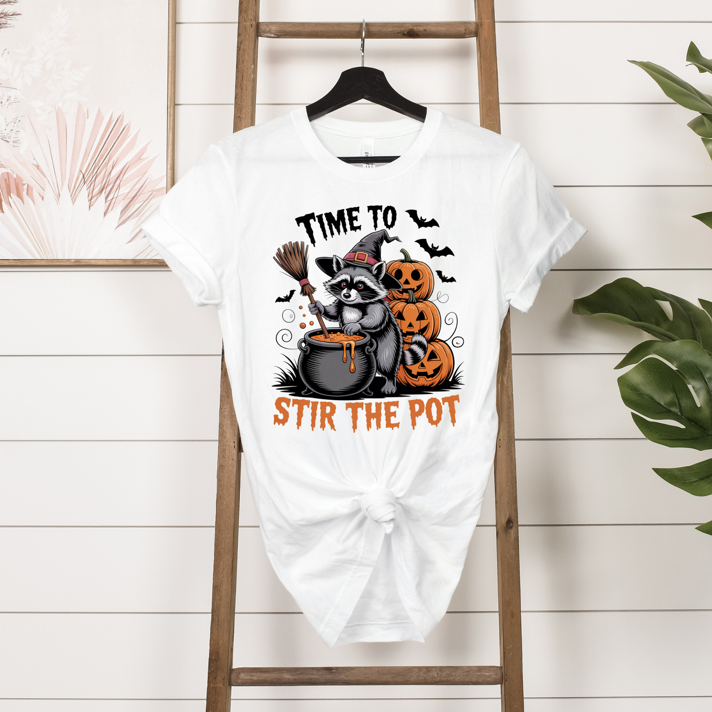 White t-shirt with Halloween-themed design featuring a raccoon, cauldron, and pumpkins on a wooden ladder.
