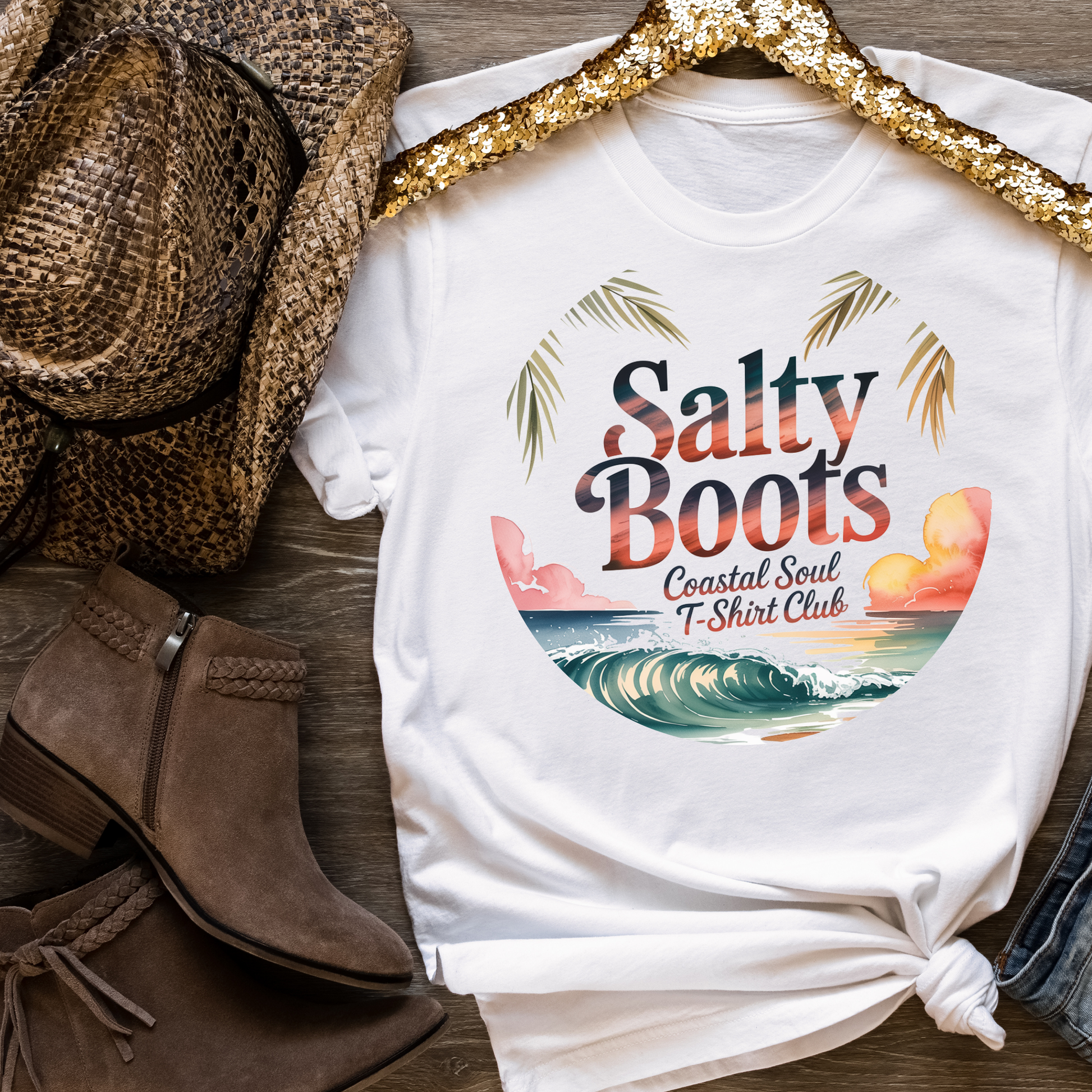 White Sunsets & Salty Boots coastal cowgirl t-shirt with beach wave design, styled with brown cowboy boots and a straw hat on wooden background