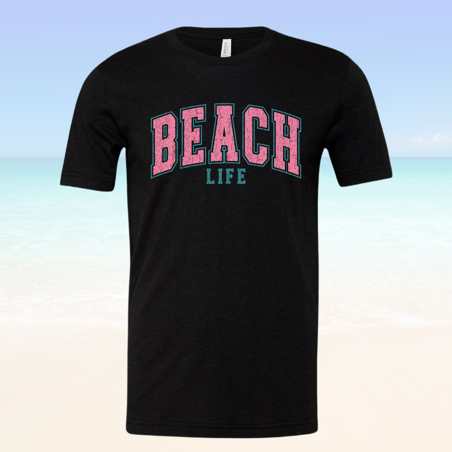 Beach Life Vibe Tee with pink and blue text on a black background, against a beach setting.