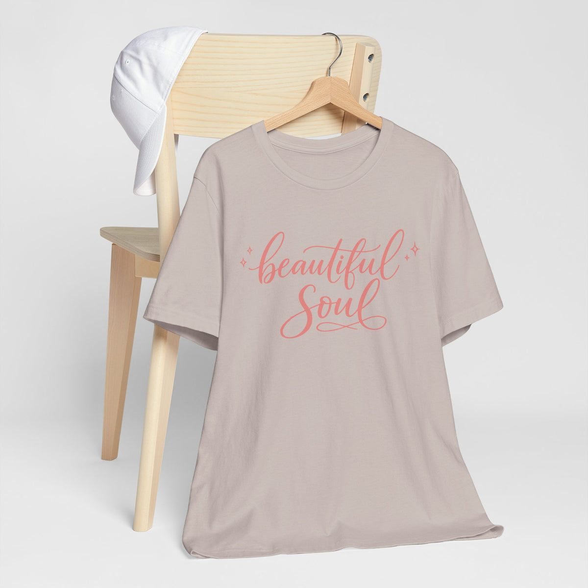 A taupe gray short-sleeve t-shirt with 'beautiful Soul' printed in coral pink script on the front, displayed on a wooden hanger draped over a light wood chair with a white towel.