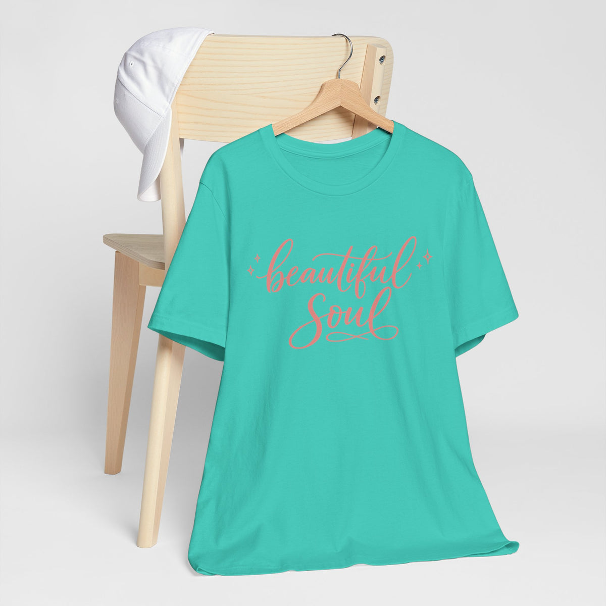 Sea Green short-sleeve t-shirt with 'beautiful Soul' printed in coral script on the chest, hanging on a wooden hanger from a light wood chair with a white towel draped over the backrest, against a clean white background.