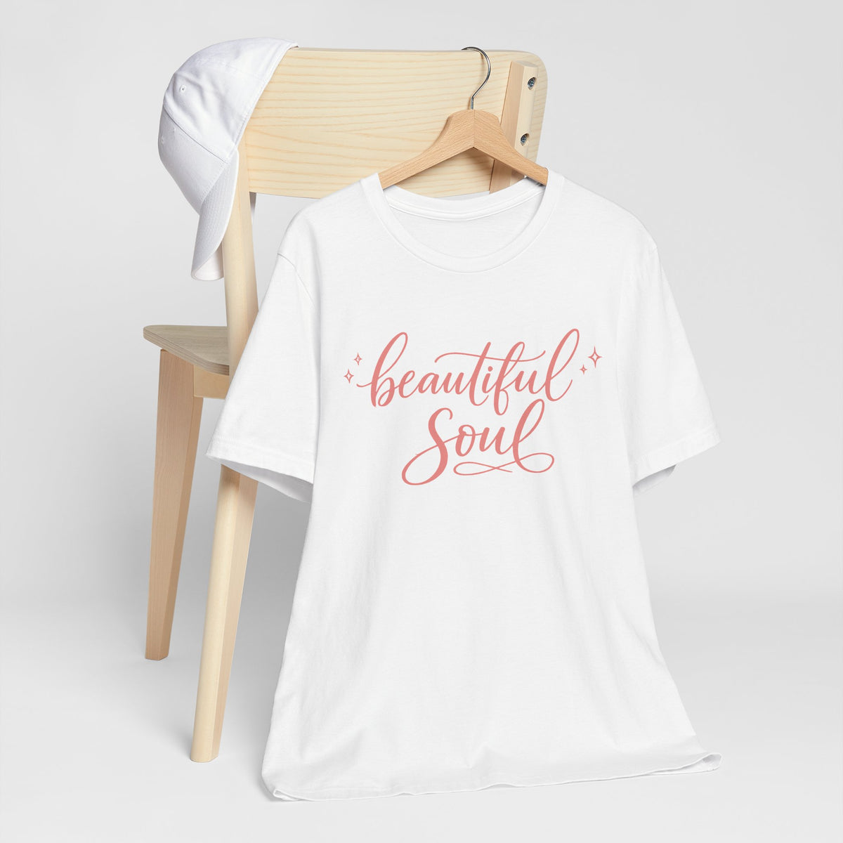 A white short-sleeve t-shirt on a wooden hanger, featuring the words 'beautiful Soul' printed in a coral pink script across the chest. The shirt is styled next to a light wood chair with a white cap hanging on its back, set against a clean, light gray studio background.