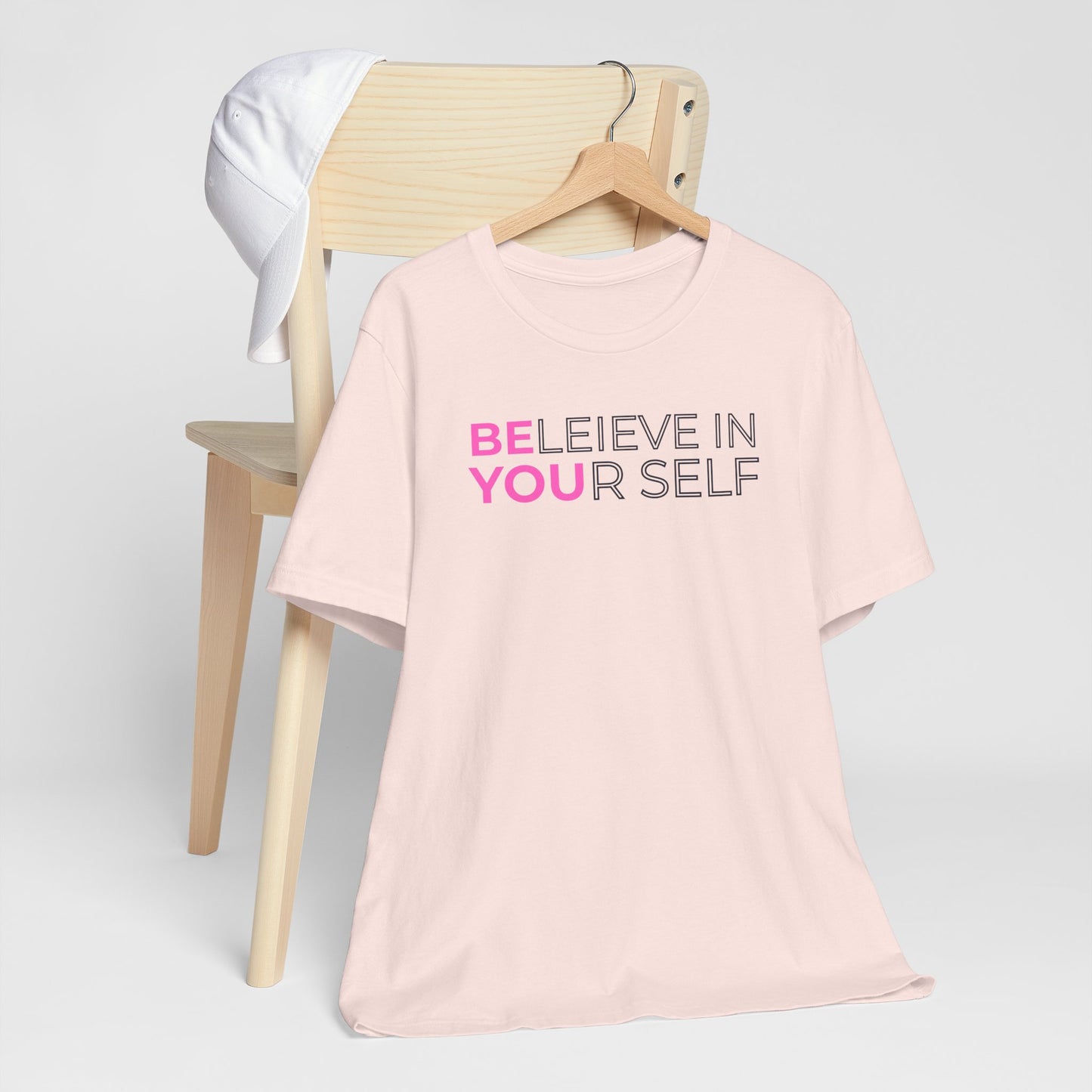 Pink t-shirt with 'BELIEVE IN YOURSELF' text on a chair against a white background