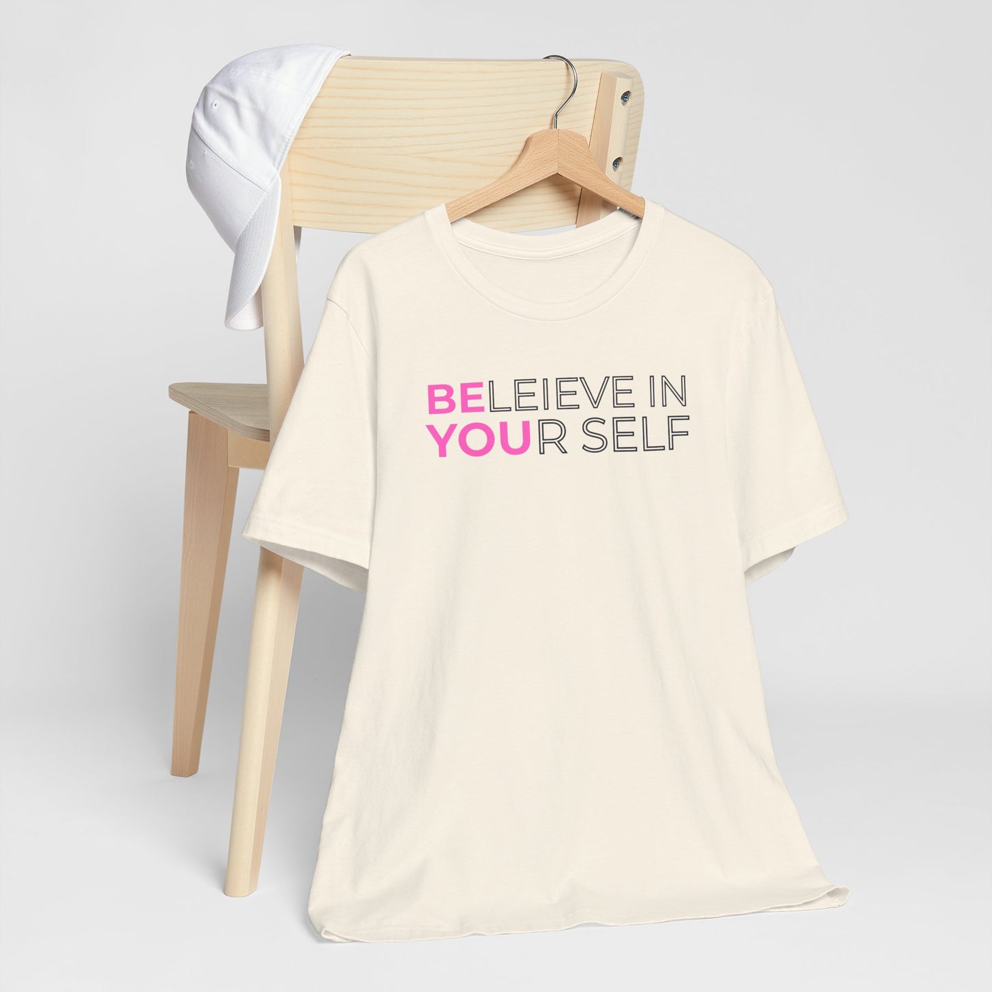 Beige t-shirt with 'BELIEVE IN YOURSELF' text on a chair against a white background