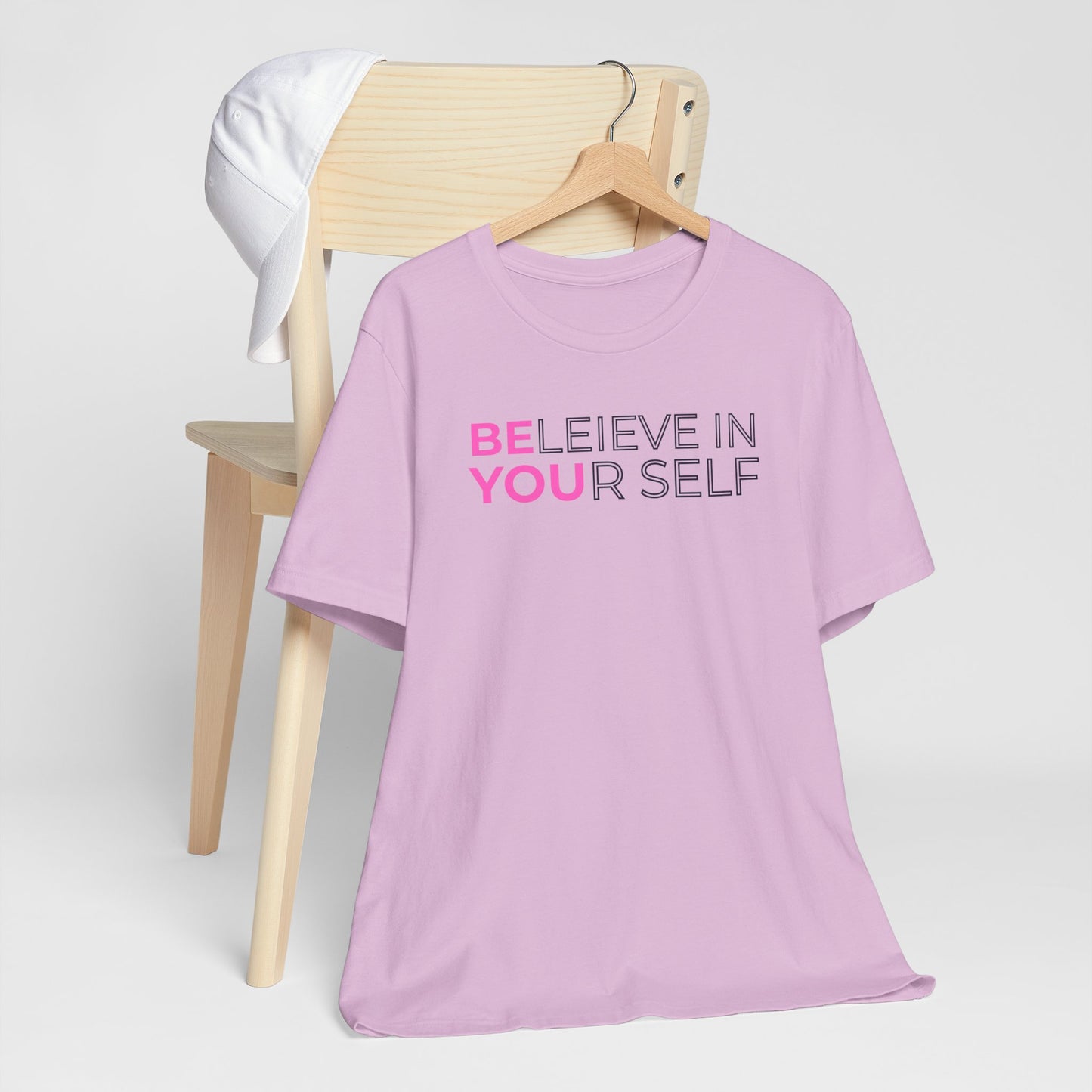 Pink t-shirt with 'BELIEVE IN YOURSELF' text on a wooden chair against a white background