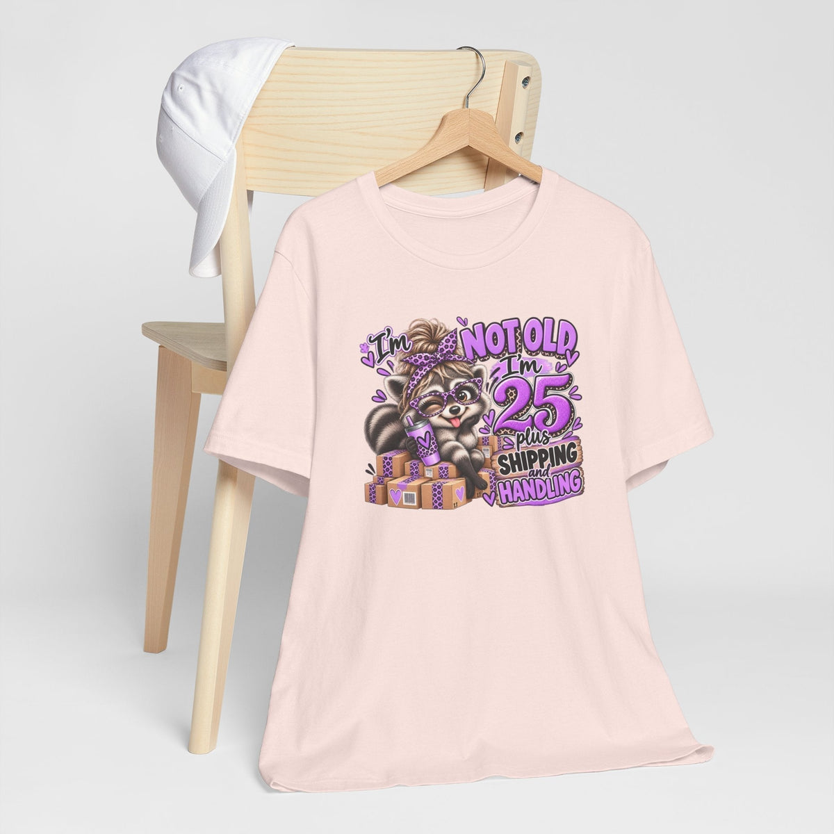 adult humor tshirt in pale pink with a colorful raccoon graphic, purple accents and the phrase “25 plus shipping and handling”