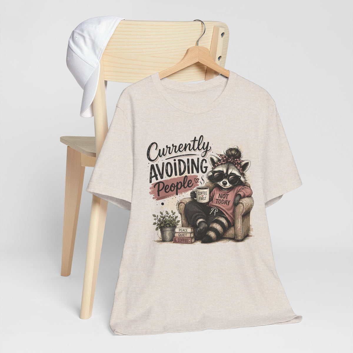 Funny tee shirt in light heather beige with a raccoon in sunglasses holding coffee beneath a humorous slogan