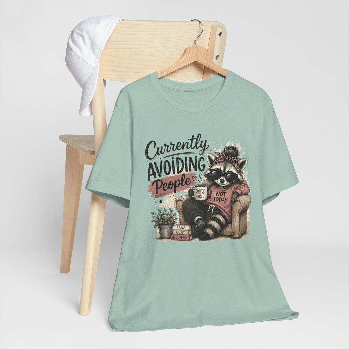 Funny tee shirt in mint green with a graphic of a sunglasses-wearing raccoon holding coffee and avoiding people