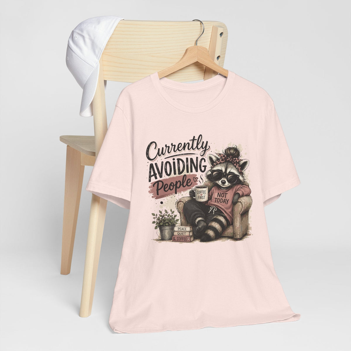 Funny tee shirt in pale pink fabric featuring a seated raccoon in sunglasses with coffee and humorous text