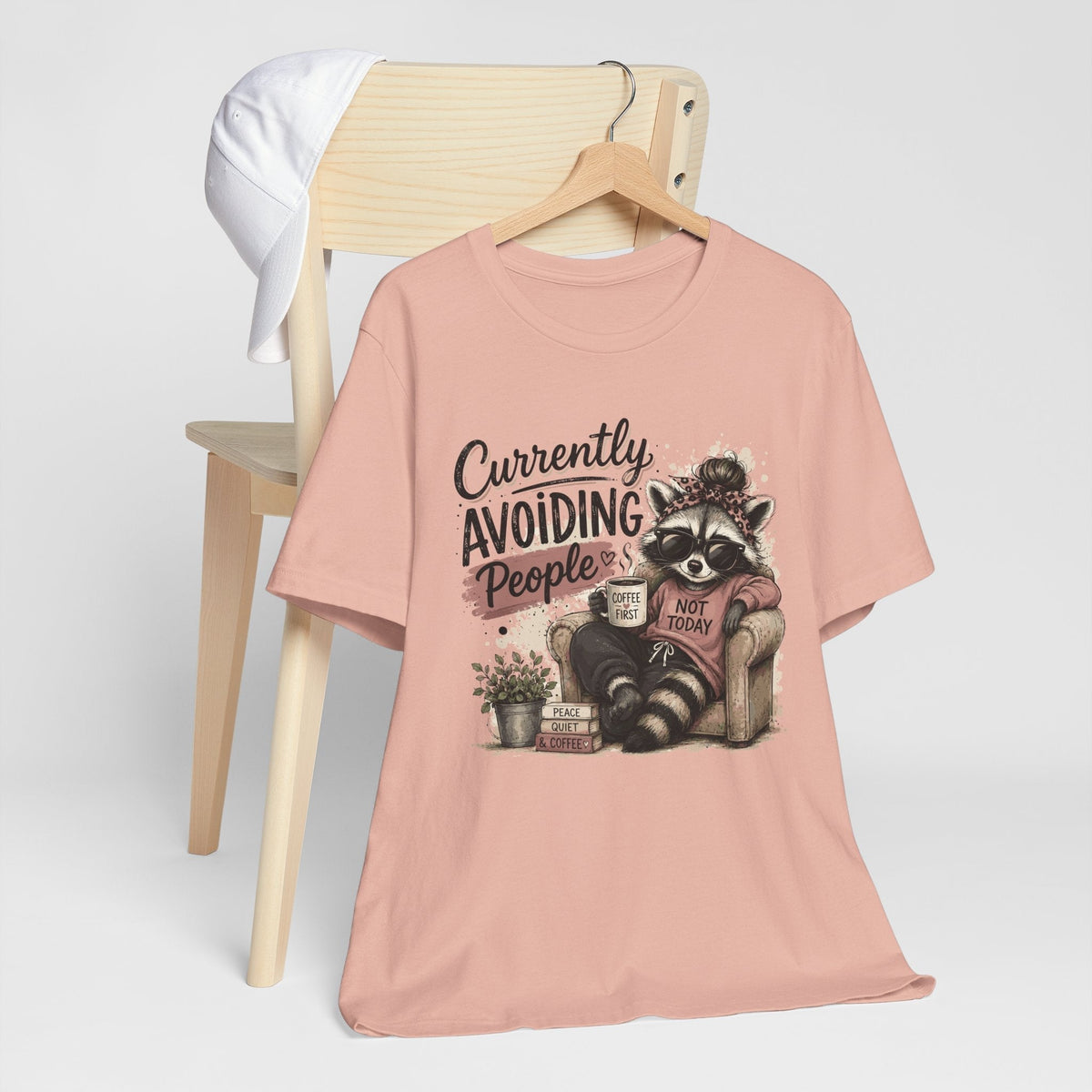 Funny tee shirt in soft pink fabric with a graphic of a sunglasses-wearing raccoon holding coffee and avoiding people