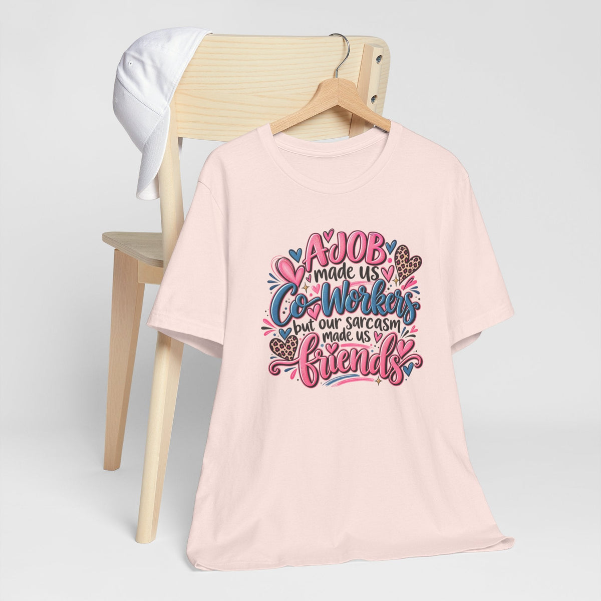 T-shirt in pale pink with colorful lettering, hearts and floral details reading “A job made us co-workers but our sarcasm made us friends,” a gift ideas for coworkers