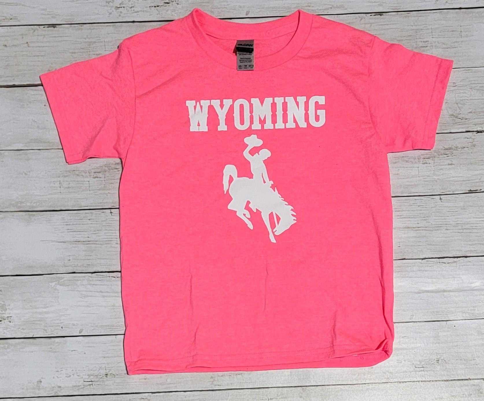 Hot pink Wyoming Cowboys youth T-shirt with white logo on wooden background.