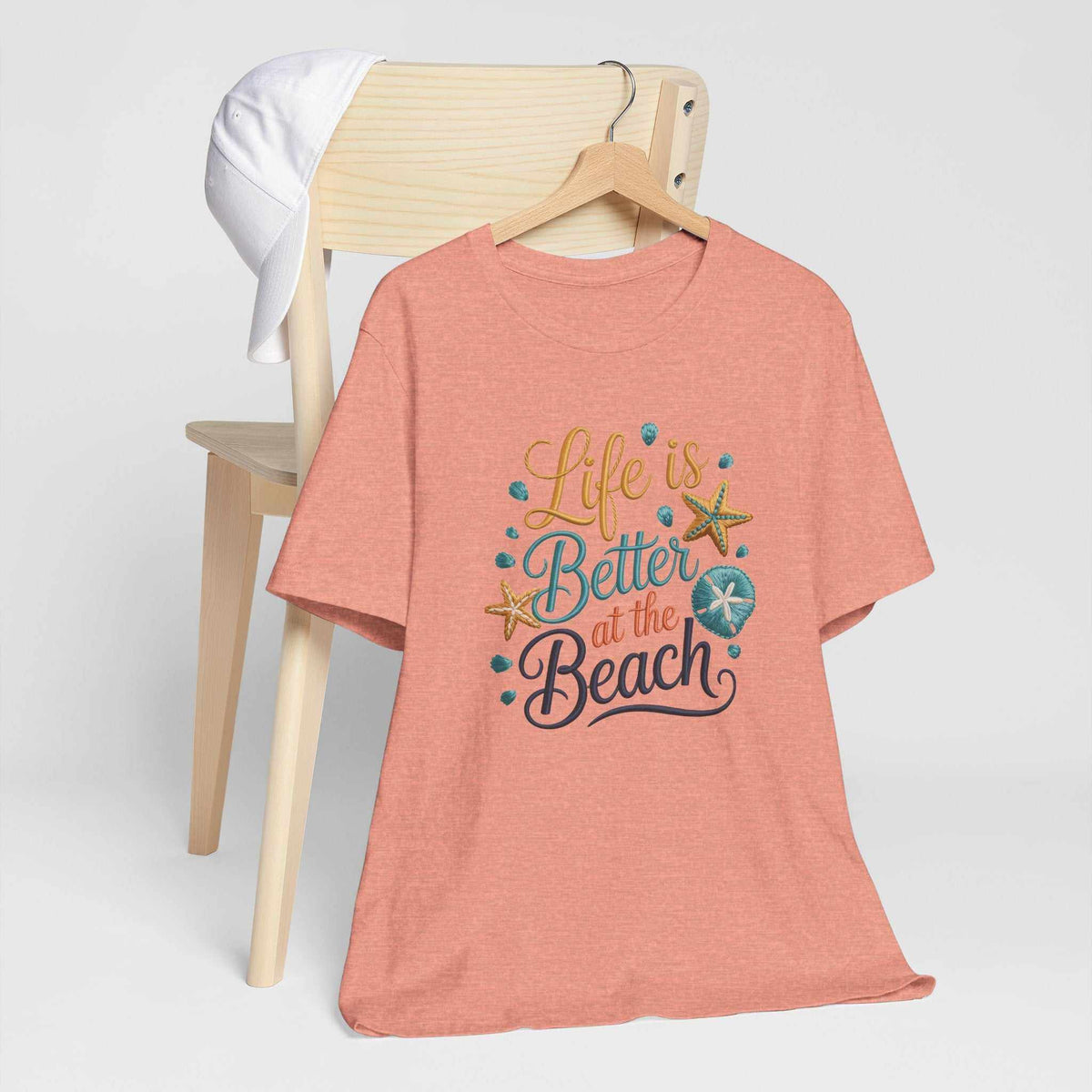 Life is better at the beach graphic T-shirt in coral with colorful lettering, seashells, and starfish