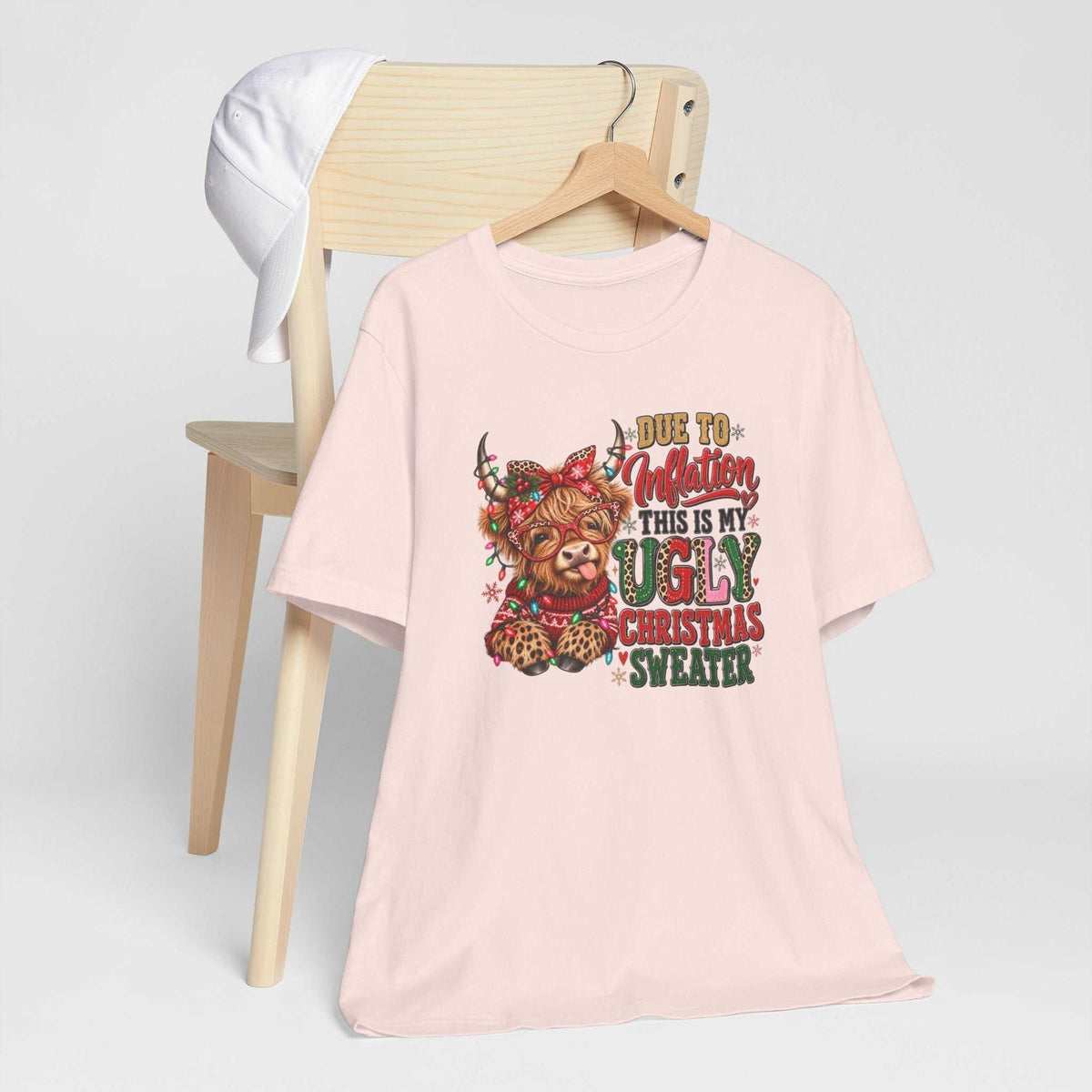 ugly christmas shirt in pale pink cotton featuring a colorful Highland cow, festive lights, leopard print and humorous text